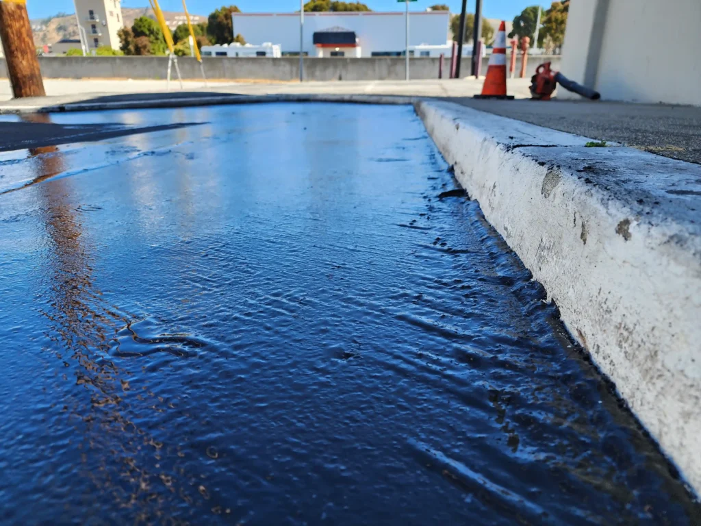 Sealcoating 2 Close-up of wet, black sealcoat reflecting the sky next to a white concrete curb. Project by We Love Paving in Northern California, CA.