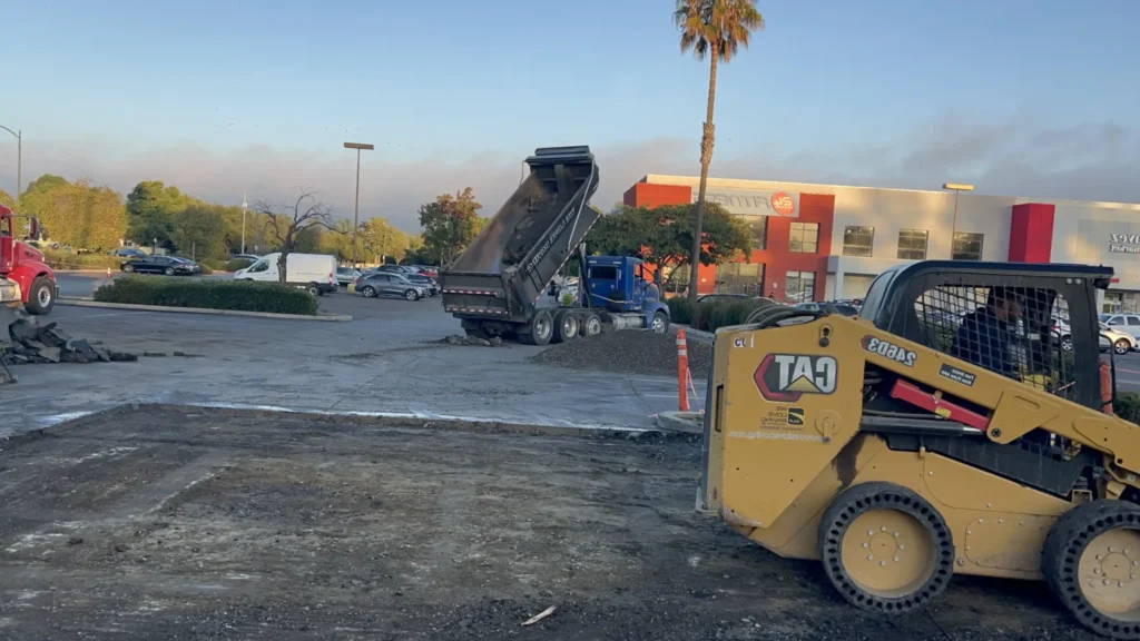 2026 Asphalt Paving Trends: Why Small Cracks Turn Into Big Problems Fast 2 A Caterpillar skid steer works while a dump truck unloads materials in a 24 Hour Fitness parking lot. Project by We Love Paving in Northern California, CA.