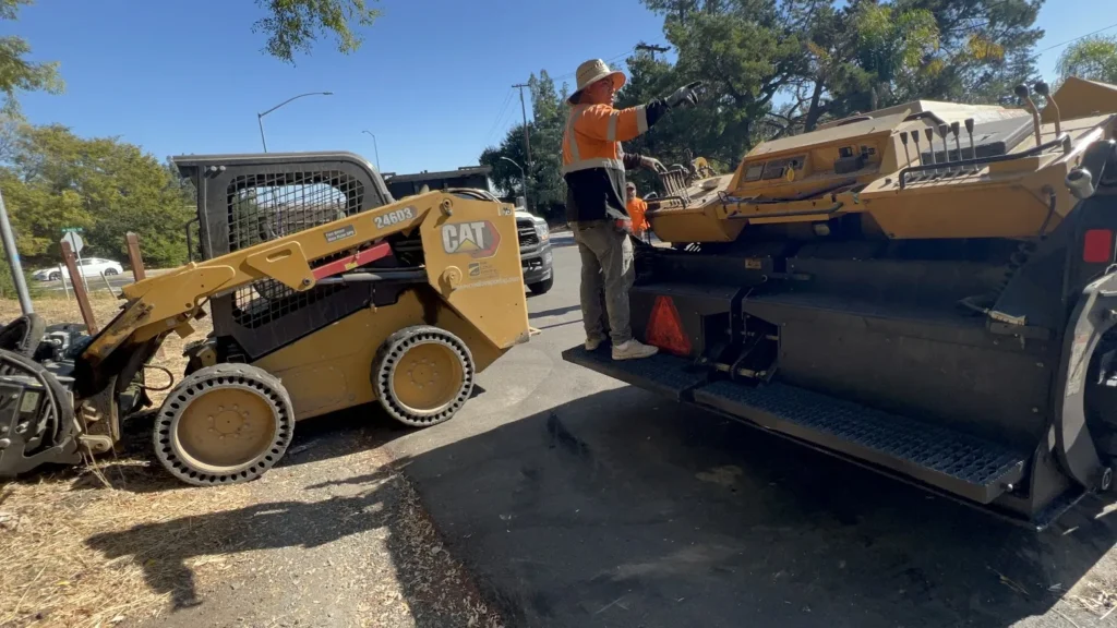 Asphalt Paving 1 A worker on a large paving machine directing a CAT skid steer loader at a job site in San Ramon. Project by We Love Paving in San Ramon, CA.