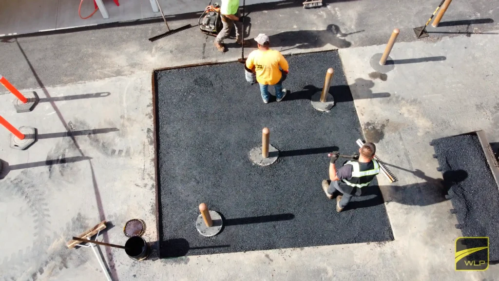 Hotel Porte Cochere Asphalt Repair 1 We Love Paving crew members using asphalt rakes to level and smooth a fresh asphalt patch in a commercial parking lot, ensuring precision around safety bollards. Project by We Love Paving in Northern California, CA.