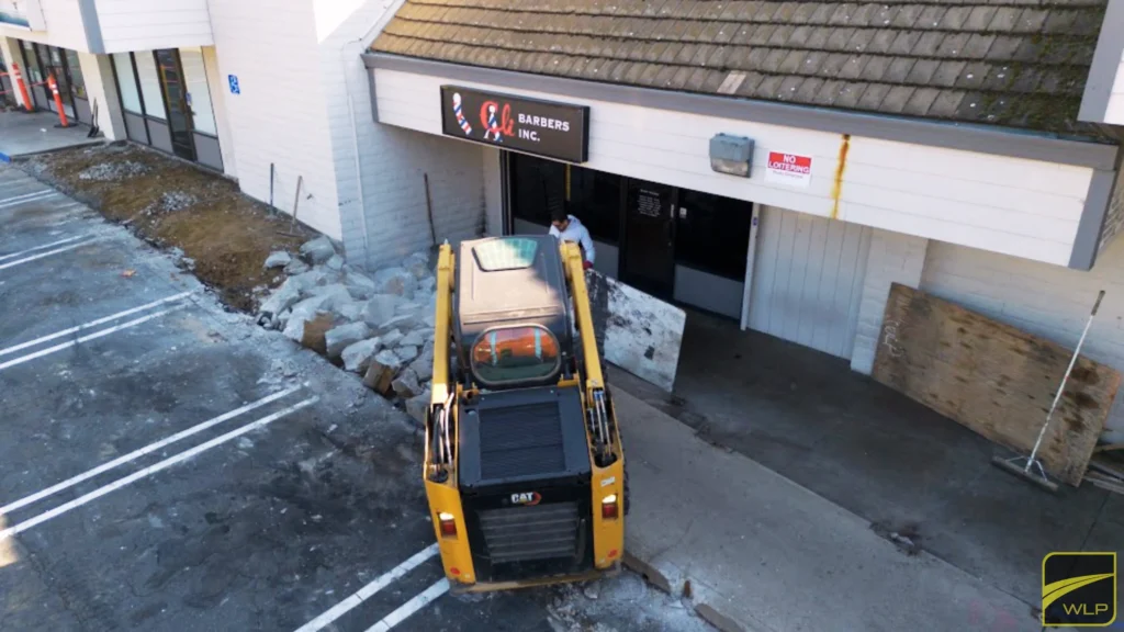 Office Park Resurfacing 1 CAT skid steer loader performing excavation work in front of Barbers Inc. storefront.