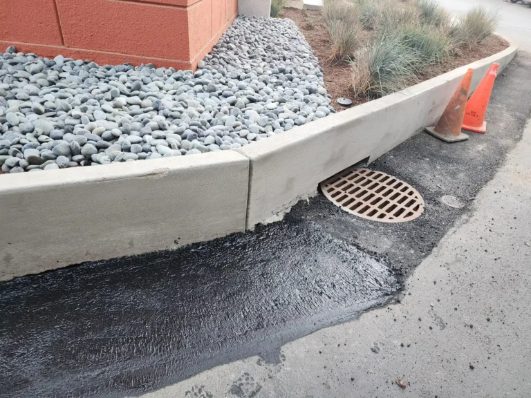 Close-up of a street storm drain with a circular grate and a curb opening, surrounded by a patch of fresh black asphalt.